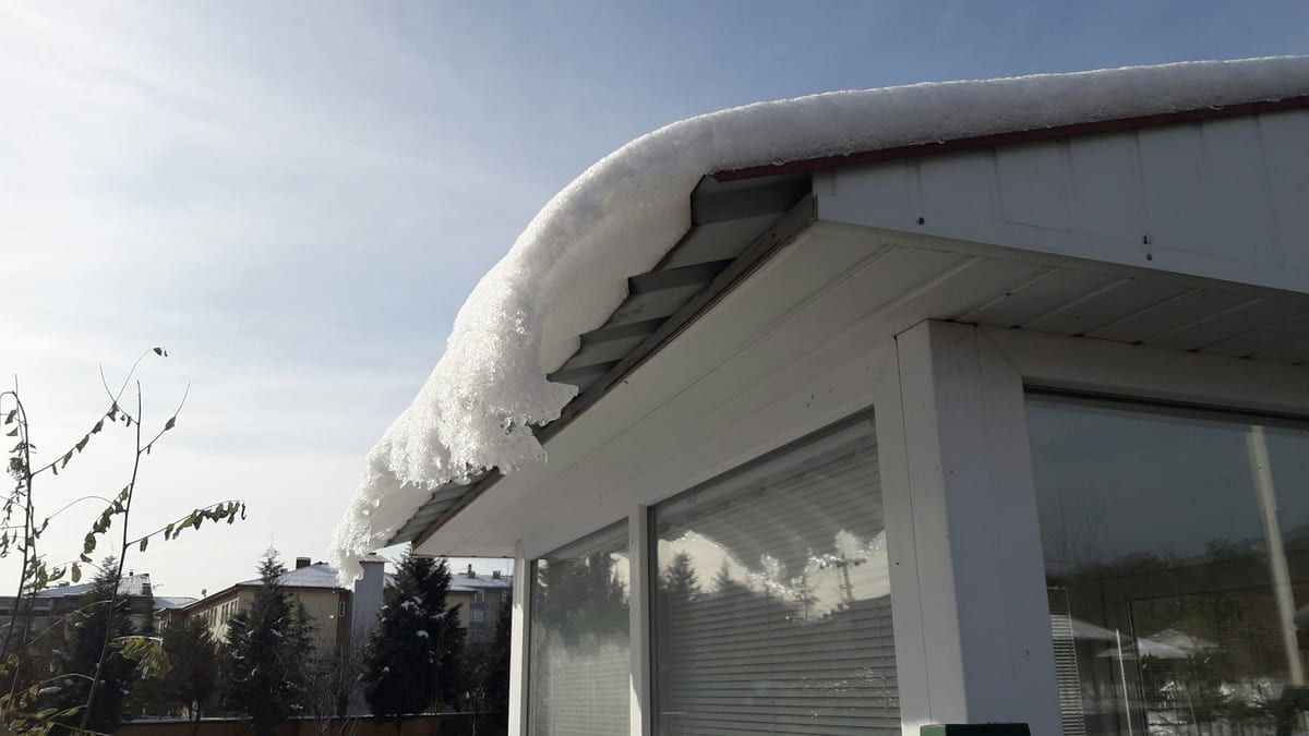 Snow on a metal roof curls over the eave above a row of windows.