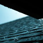 Rainwater runs off the edge of an asphalt shingle roof and drips from the lower roof edge.