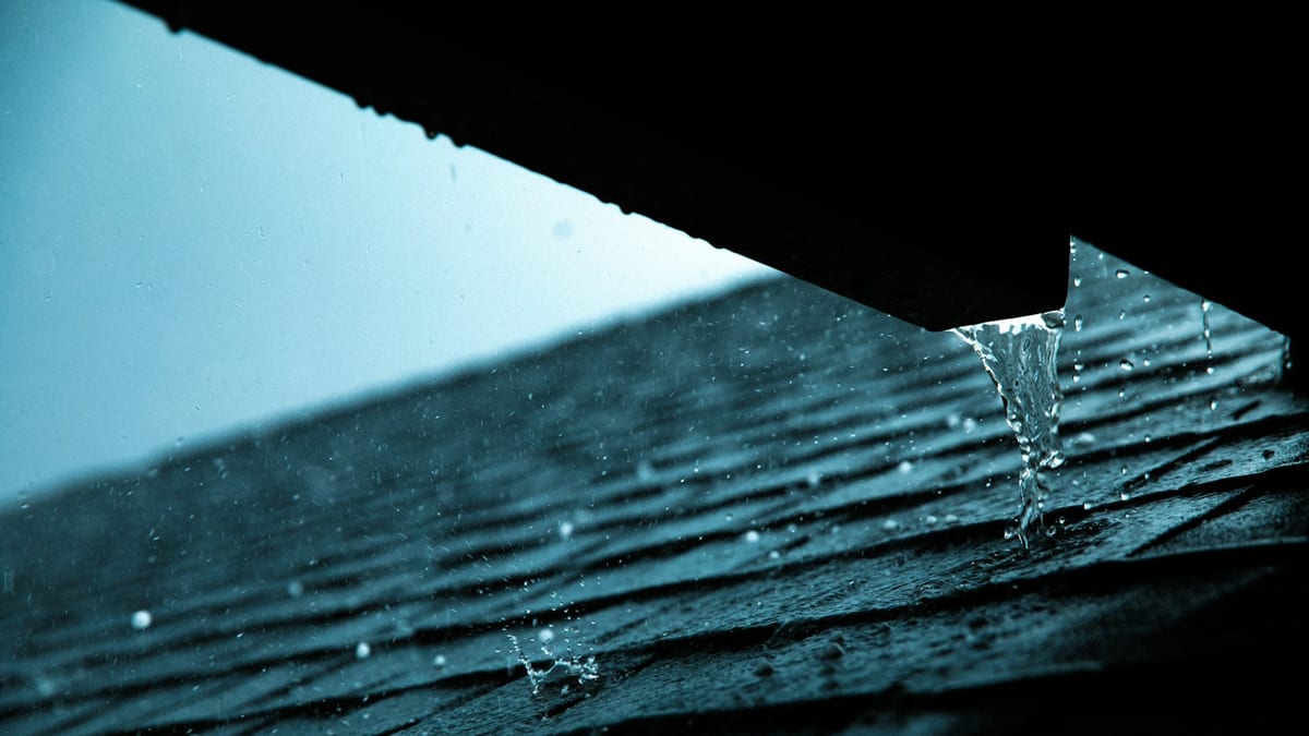 Rainwater runs off the edge of an asphalt shingle roof and drips from the lower roof edge.