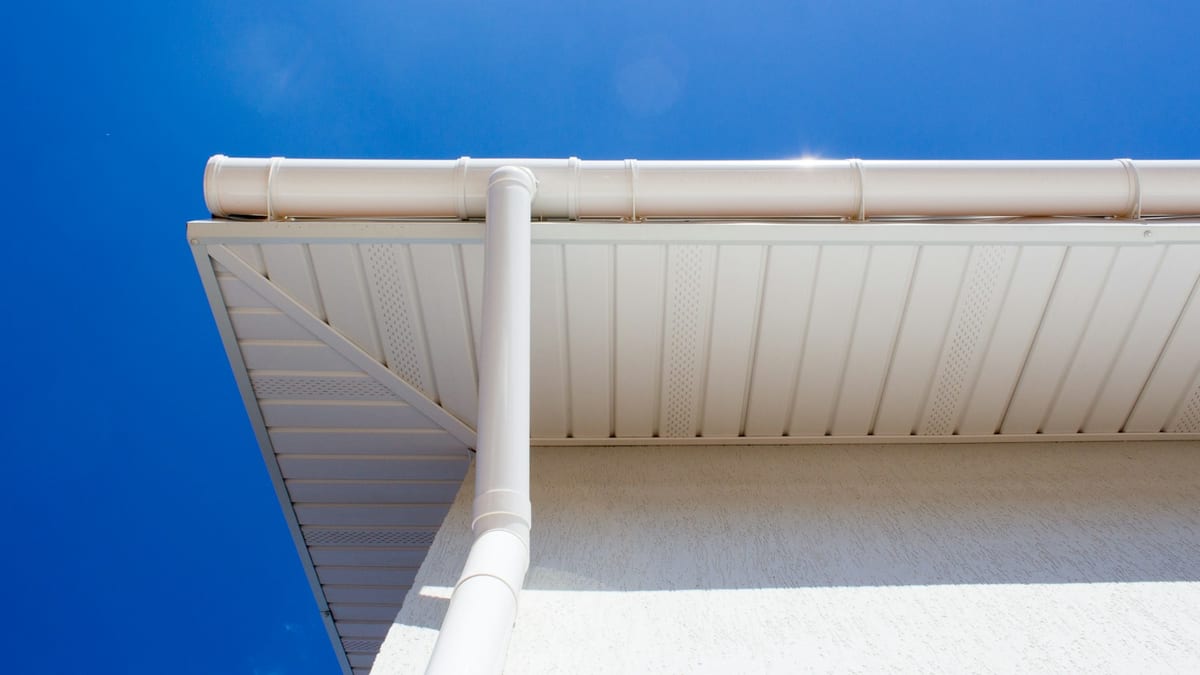 White eavestrough and downspout along the roof edge, seen from below against a clear blue sky.