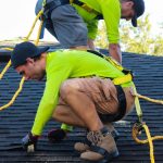 Roofer in a safety harness crouches on a dark shingle roof and works on the shingles with gloved hands.